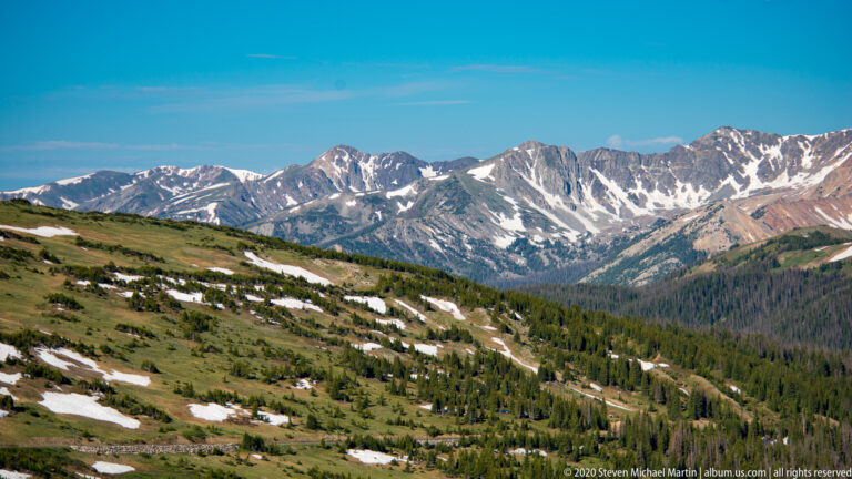 A mountain range with snow overlooks grassy slopes and evergreen trees. in the alpine landscape.