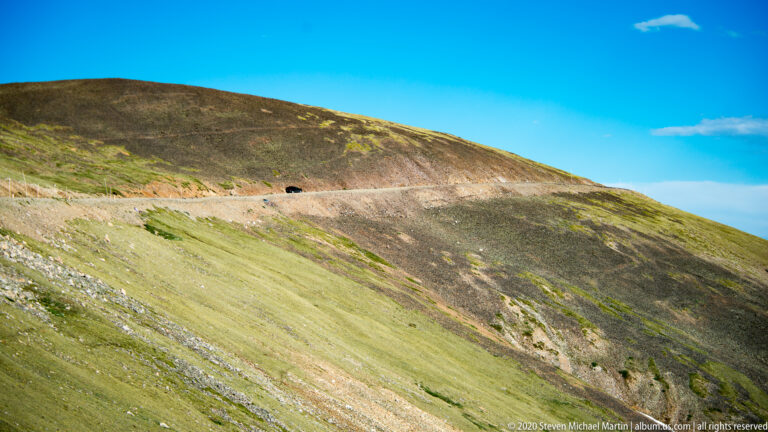 A narrow road crosses an open grassy alpine slope beneath a clear sky.