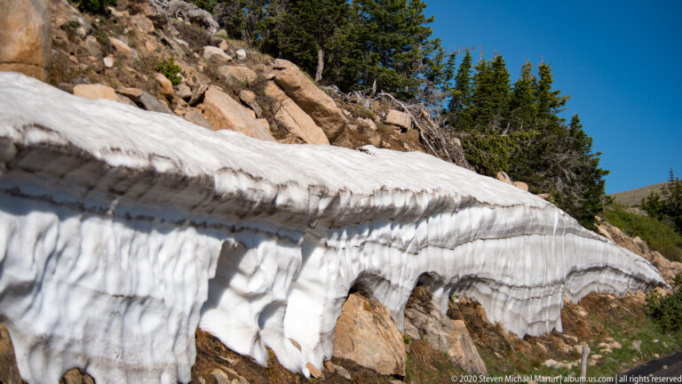 A deep snowbank overhangs exposed rocks beneath conifers and blue sky. in the alpine landscape.