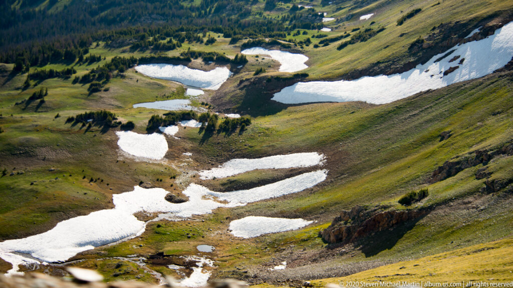 Irregular snow patches lie in shallow drainage folds across a green alpine basin.