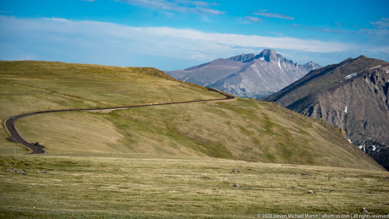 A curving paved road crosses alpine grassland toward a rugged snow-patched peak.