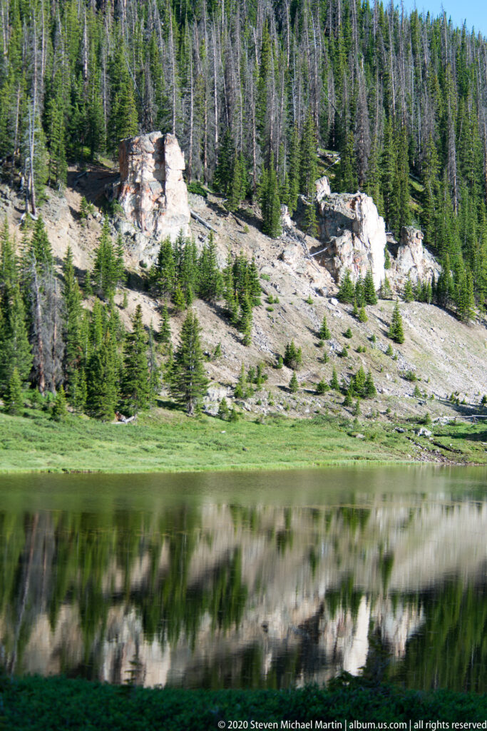 A still lake mirrors conifers and pale rock outcrops beside a green alpine meadow.