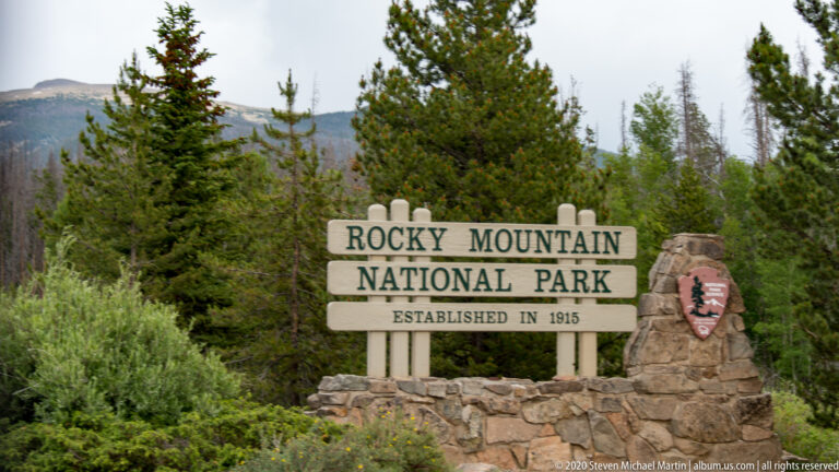 A wooden sign names Rocky Mountain National Park and its 1915 establishment beside a stone wall.