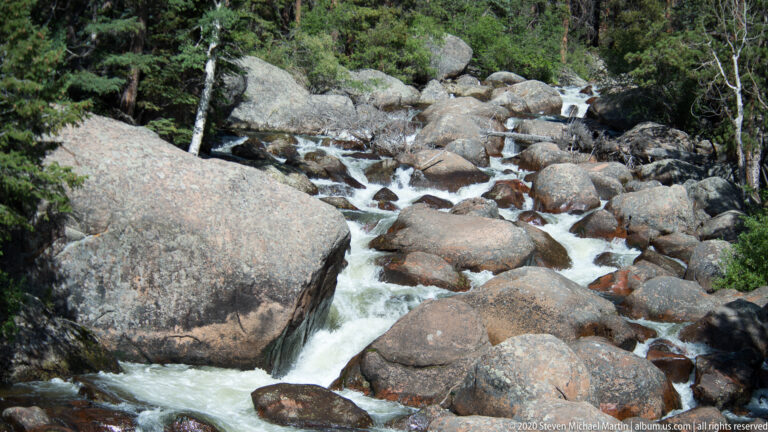 A rocky creek cascades between large boulders through a shaded, forested mountain channel.