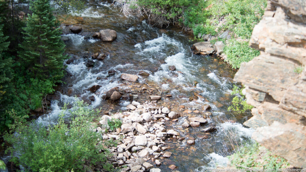 An elevated view looks down on a rocky creek winding between boulders, shrubs, and trees.
