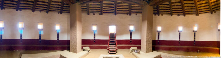 Timber roof structure and tall supports dominate a view toward the far stairway inside the reconstructed Great Kiva.