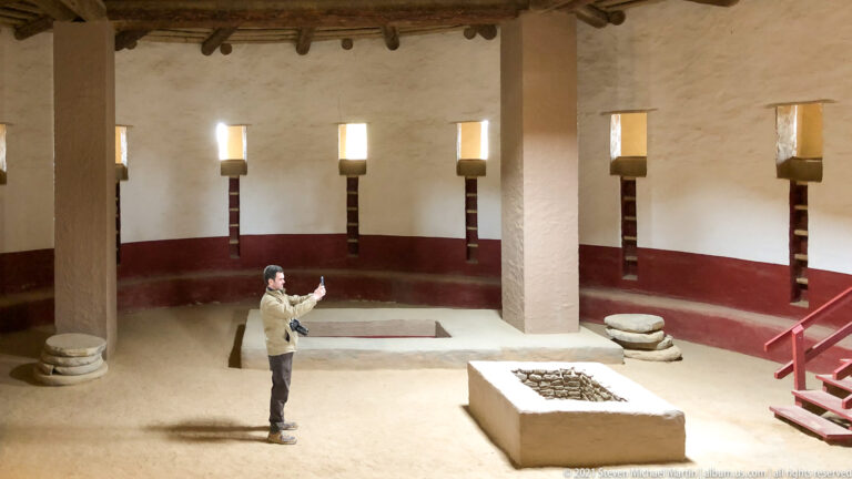 Visitor raises a camera inside the reconstructed Great Kiva as a long shadow crosses the floor.
