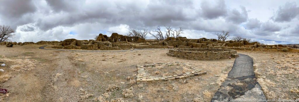 Wide archaeological landscape with low masonry ruins, circular stone features, and a paved visitor path beneath cloudy sky.