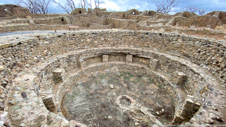 Overhead view into a circular excavated kiva with concentric stone walls, wall recesses, and a small central feature.