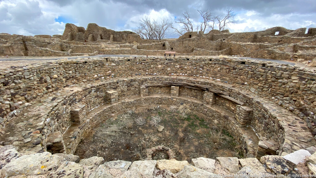 Wide elevated view of a circular excavated kiva with extensive masonry ruins rising behind it.