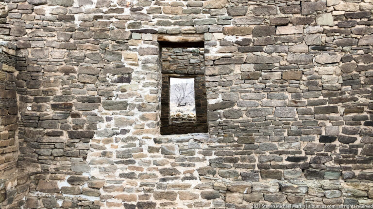 Standing stone wall with two vertically aligned rectangular openings framing bare trees beyond.