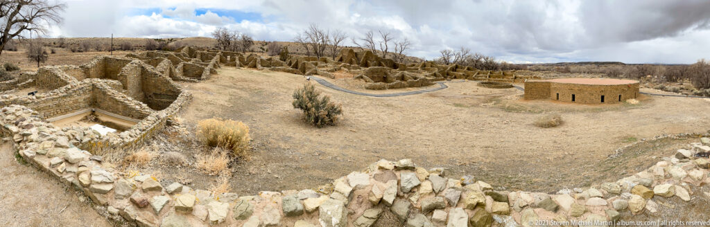 Elevated wide view over excavated room blocks toward the large circular reconstructed Great Kiva at right.