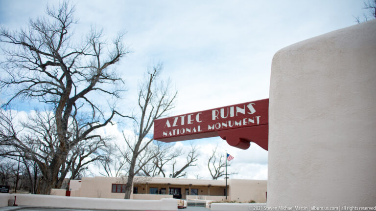 Red Aztec Ruins National Monument entrance sign mounted beside a white stucco pier with bare trees behind.