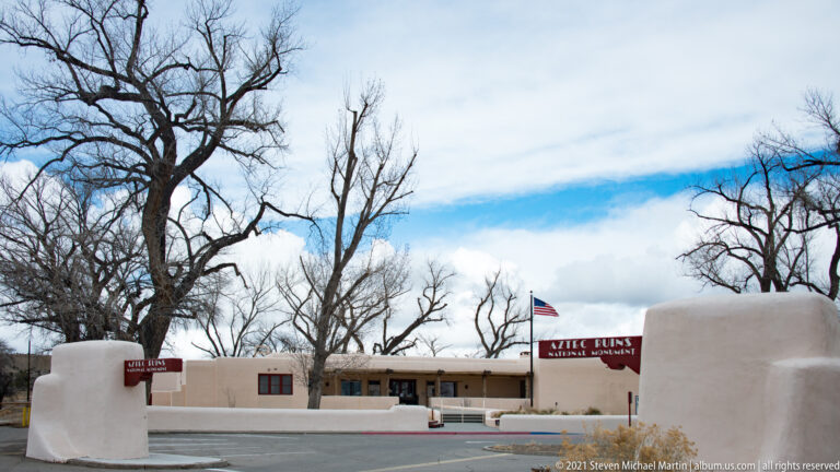Wide exterior of Aztec Ruins National Monument visitor center with white stucco entrance walls, red sign, flag, and bare trees.