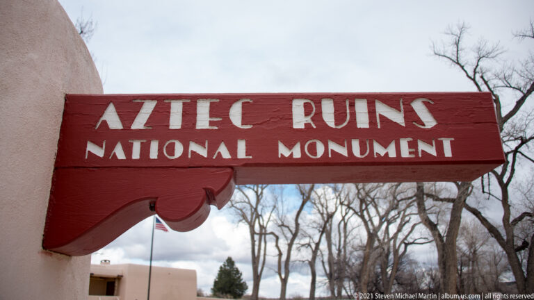 Close view of red wooden Aztec Ruins National Monument sign with white lettering against bare trees and cloudy sky.