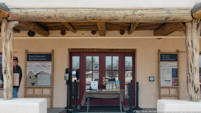 Visitor center entrance with red-framed doors, interpretive panels, table, and heavy natural-log porch supports.