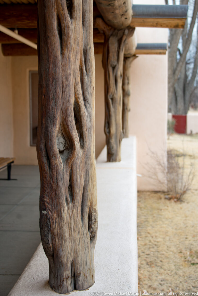 Close view of a thick gnarled natural-log porch post on a white stucco base, with similar posts receding behind.