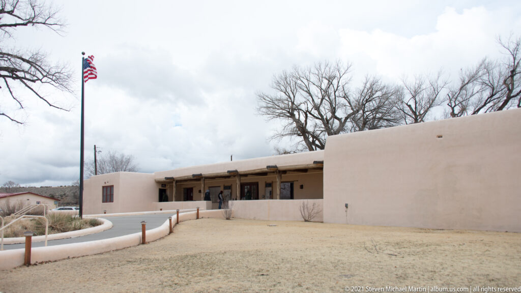 Curving walkway approaches the low stucco visitor center beneath an American flag and leafless trees.