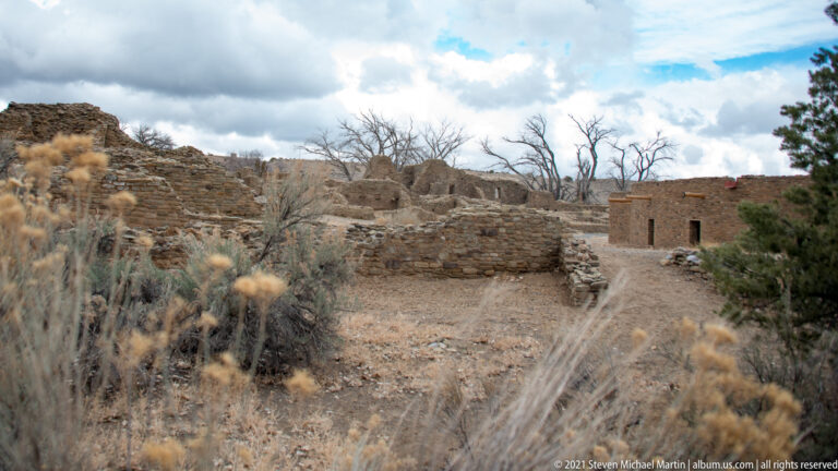 Low stone room walls and taller ruins seen through dry winter plants beneath a cloudy sky.