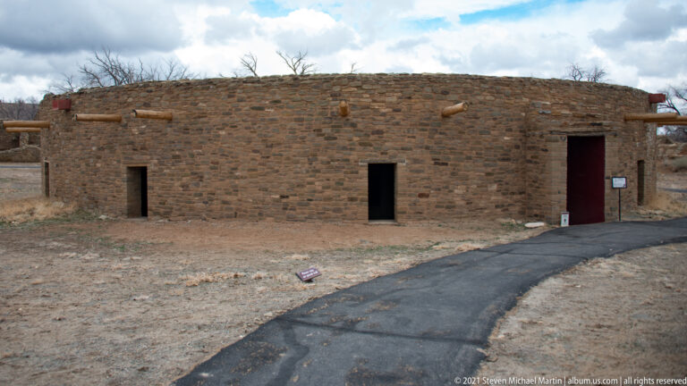 Curved stone exterior of the reconstructed Great Kiva beside a paved path, with small openings and projecting roof timbers.