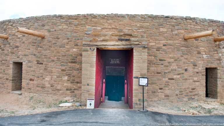 Red-painted entrance centered in the curved stone wall of the reconstructed Great Kiva, with signs and projecting timbers.