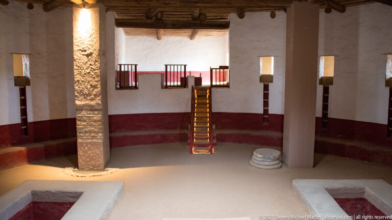 Ladder rises between large roof supports inside the reconstructed Great Kiva, with floor recesses in the foreground.