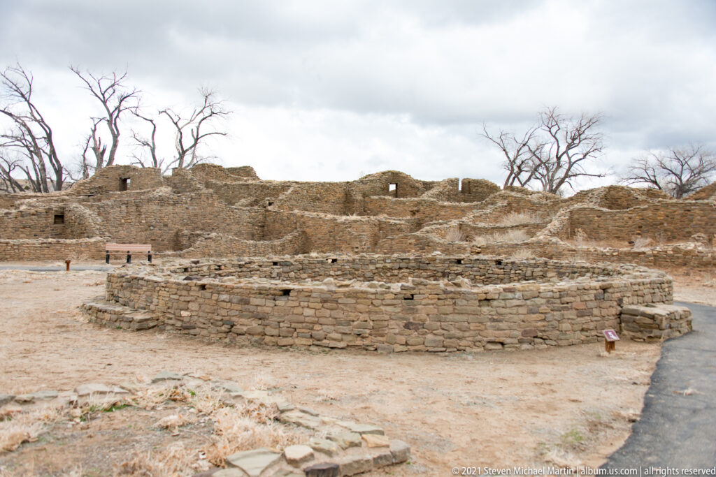 Circular excavated kiva in front of extensive stone room ruins, with a paved visitor path at right.