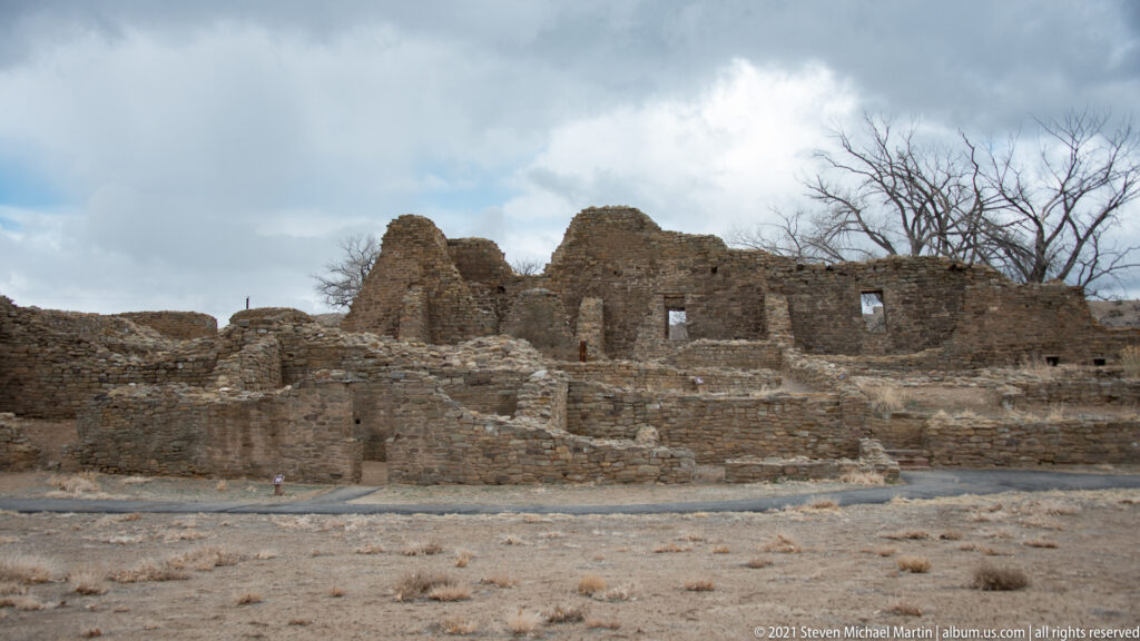 Multi-level stone ruin walls with rectangular openings rise behind lower excavated rooms under a cloudy winter sky.