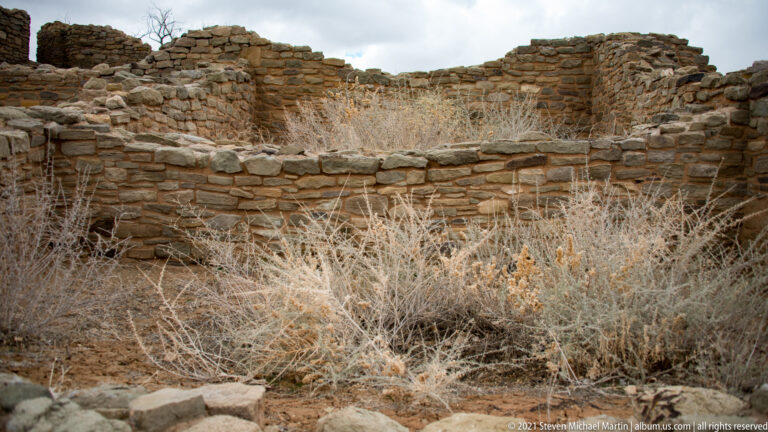 Dry shrubs fill a low stone-walled room, with higher masonry ruins behind.