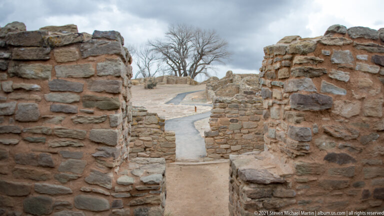 Narrow opening through thick stone room walls frames a paved visitor path and low ruins outside.