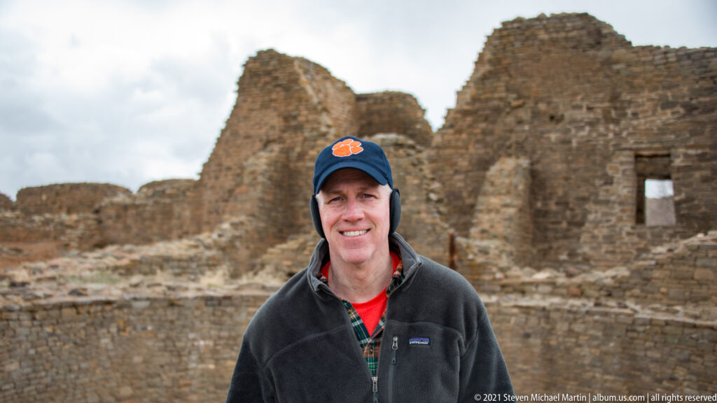 Smiling visitor in dark jacket and cap stands before tall stone ruin walls at Aztec Ruins.