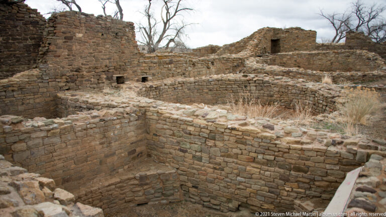 Elevated view into deep rectangular stone rooms adjoining curved masonry walls and higher ruins.