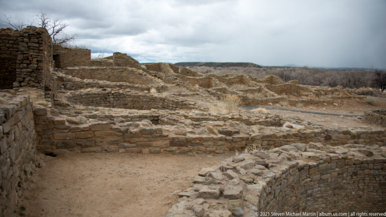 Elevated view across excavated stone rooms toward leafless woodland and distant terrain beneath a cloudy sky.