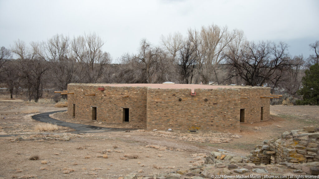 Elevated view of the reconstructed Great Kiva as a low circular stone building with dark openings and projecting timbers.