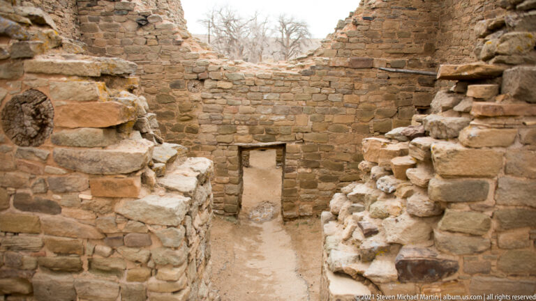 Narrow stone corridor leads to a low rectangular doorway beneath broken masonry walls.