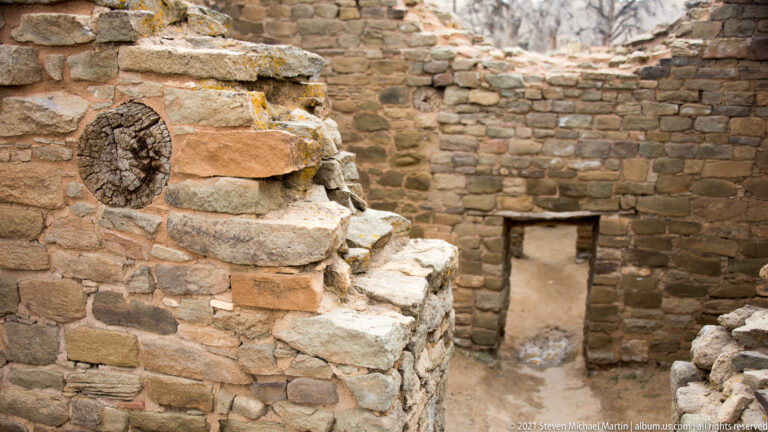 Cracked circular timber end embedded in a stone wall beside a narrow passage and doorway.