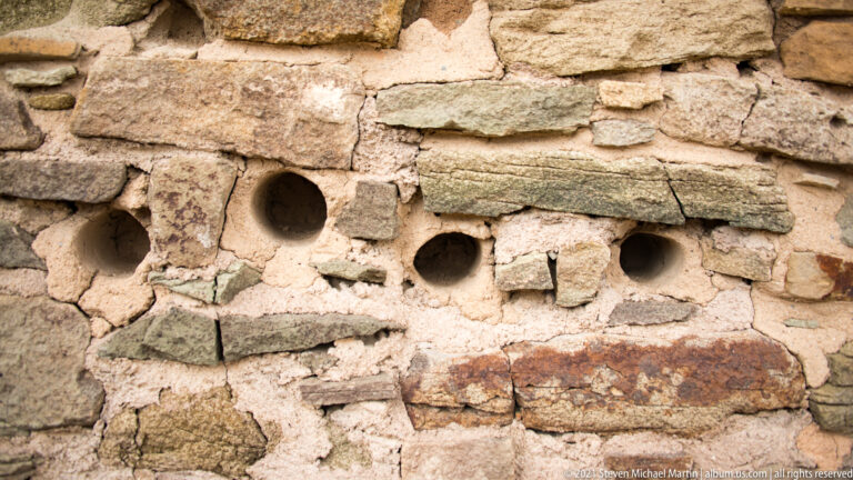 Four small round holes form a horizontal row within pale mortar and irregular stone masonry.