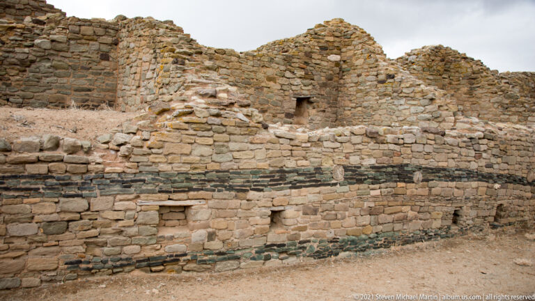 Curved tan masonry wall crossed by dark horizontal stone bands, with small openings and higher ruins behind.