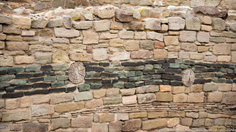 Close patterned stone wall with dark horizontal courses and two circular weathered timber ends.