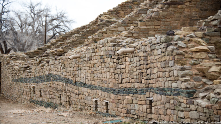 Long pale stone wall with a narrow dark masonry band receding toward taller broken ruins.