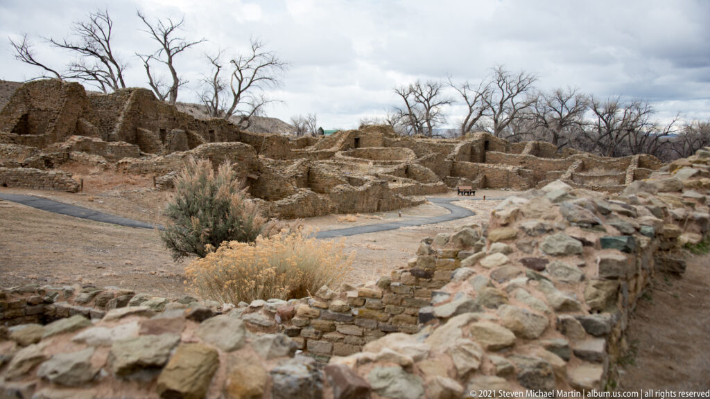 Curving paved visitor path winds among low stone room ruins, dry shrubs, and taller walls beneath leafless trees.