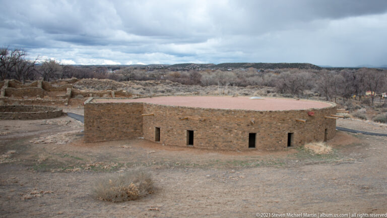 Elevated view of the circular reconstructed Great Kiva with excavated ruins, woodland, and distant terrain behind.
