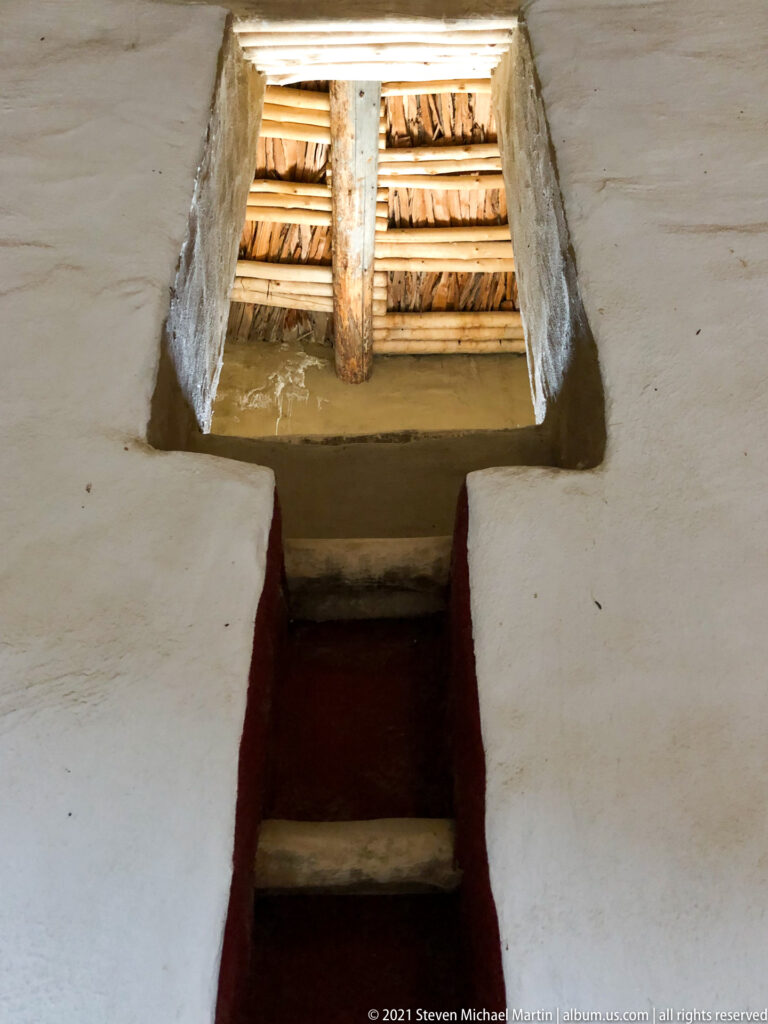 Short stairway rises through a red-lined opening toward the timber roof of the reconstructed Great Kiva.