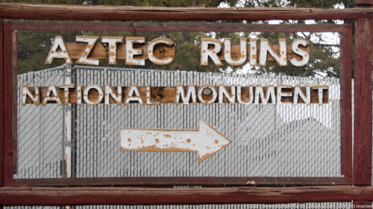 Weathered red metal Aztec Ruins National Monument sign with raised lettering and a large white arrow pointing right.