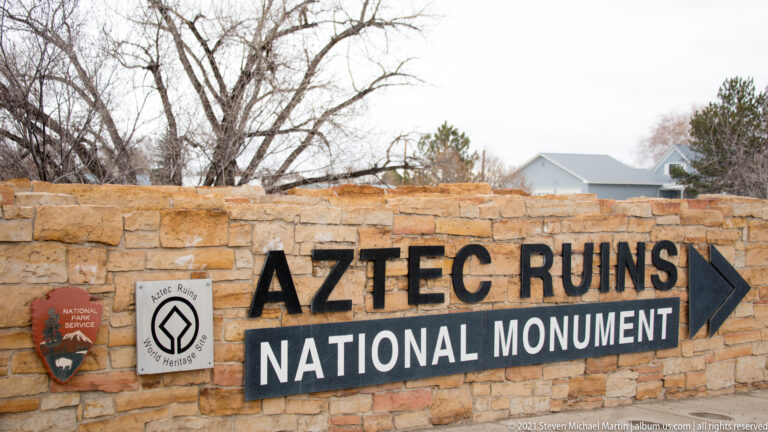 Stone entrance wall reading Aztec Ruins National Monument with right arrow, National Park Service arrowhead, and World Heritage emblem.