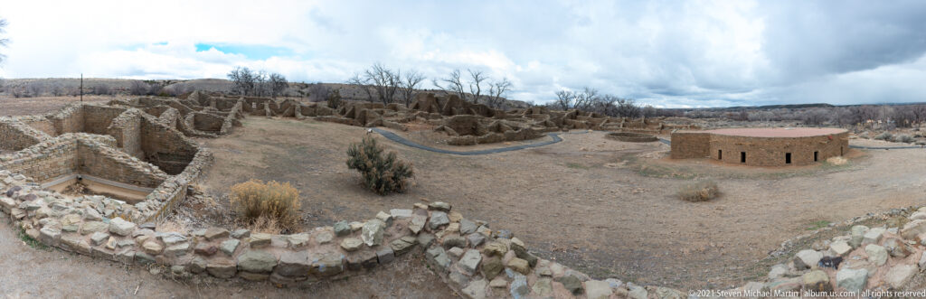 Wide elevated panorama of excavated stone room blocks and the circular reconstructed Great Kiva beneath a dramatic winter sky.
