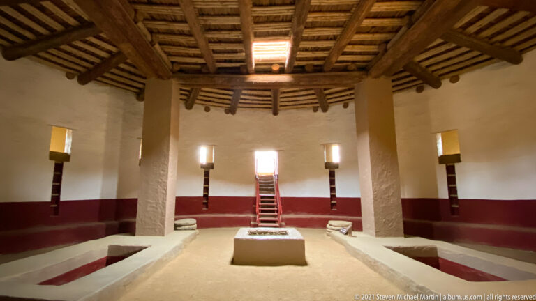 Elevated centered view across the reconstructed Great Kiva with supports, central feature, floor recesses, and far stairway.