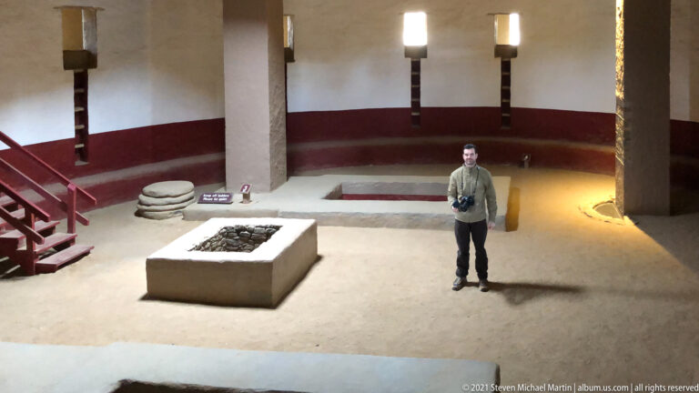 Visitor stands inside the reconstructed Great Kiva beside its central masonry feature and large roof supports.