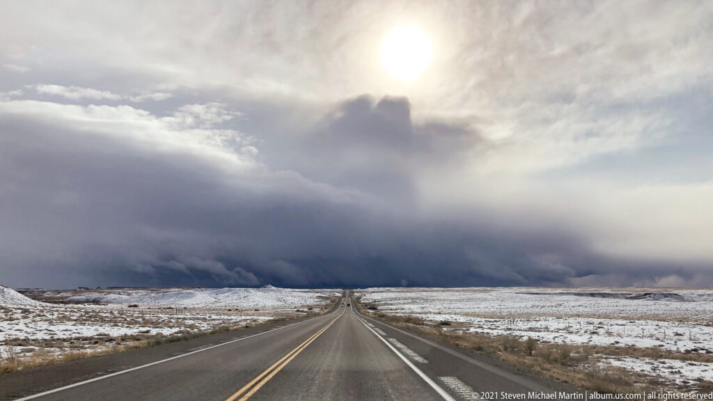 A straight highway crosses a snowy plateau toward a dark storm, with sunlight breaking through clouds overhead.