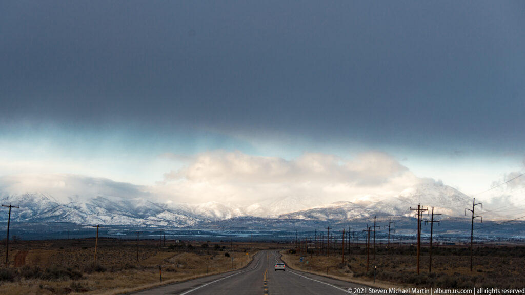 A highway and utility poles lead toward a distant snow-covered mountain range beneath low clouds.